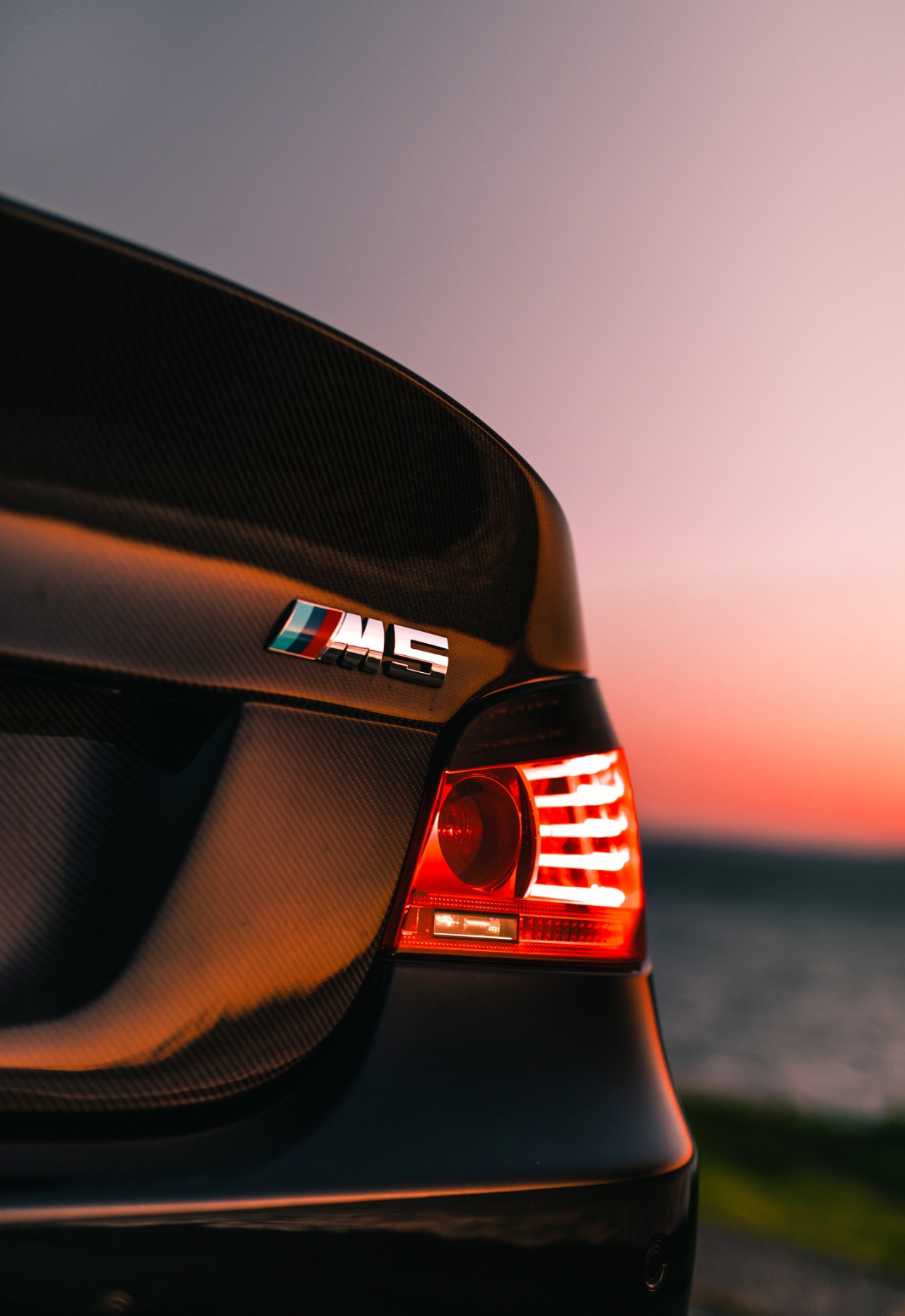 BMW retrofitting with parts from higher versions – rear lights and details from the M series Tył sportowego BMW z emblematem M5, elementami z włókna węglowego i zmodernizowanymi tylnymi światłami LED na tle zachodzącego słońca.